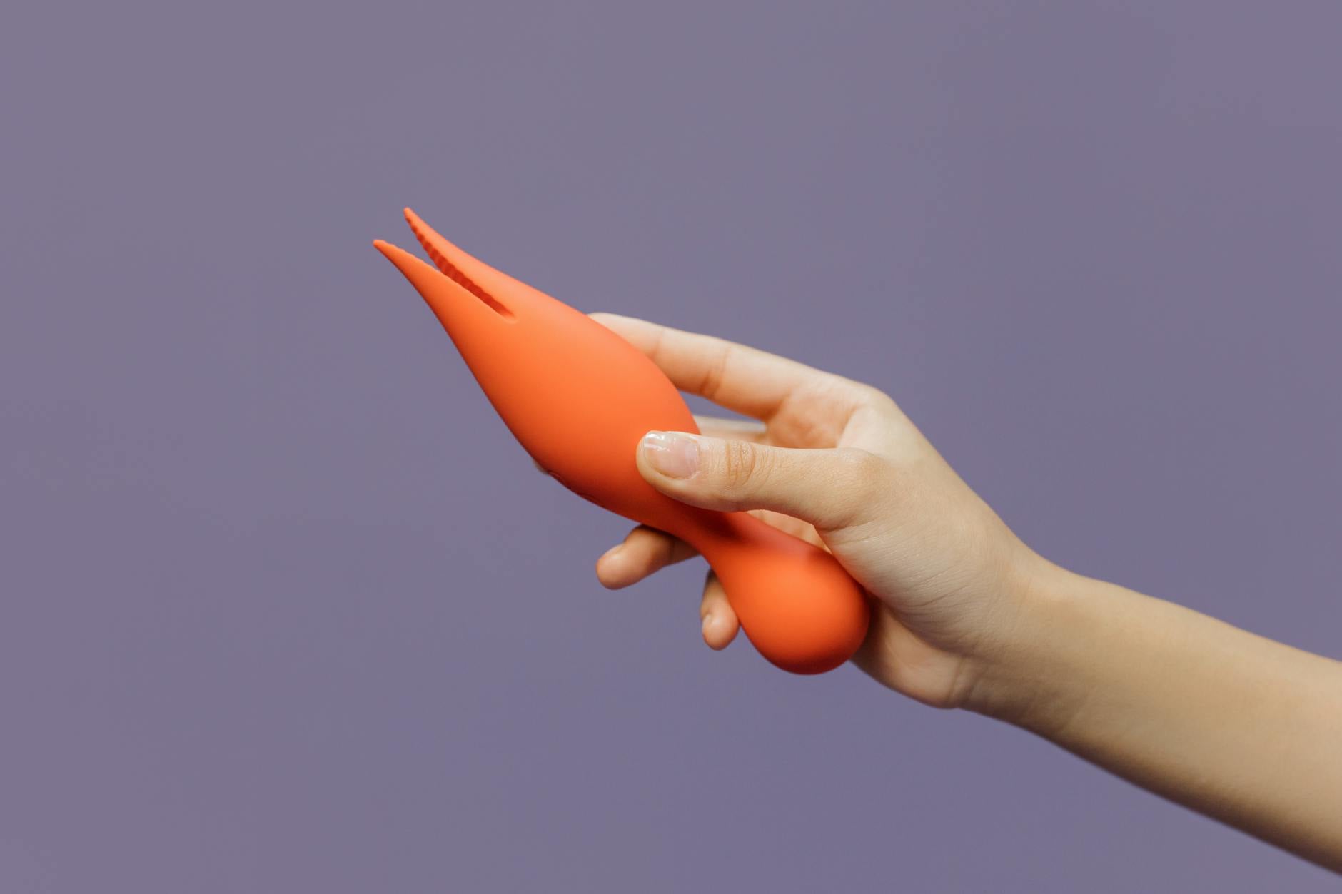 Mano sosteniendo un vibrador de color naranja contra un fondo morado minimalista, mostrando sensualidad moderna